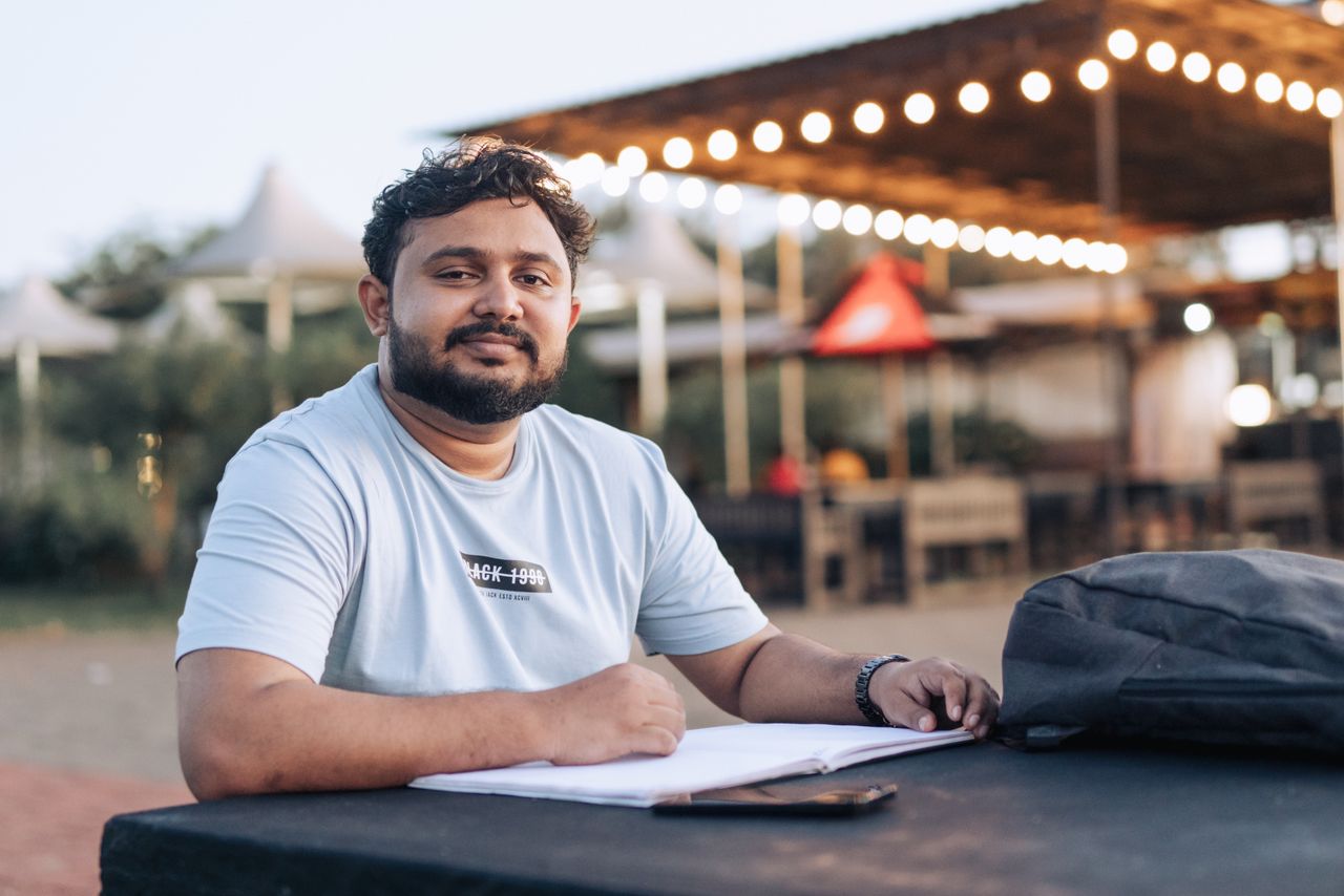 A man with a beard studying outside under string lights, with a notebook and backpack on the table.