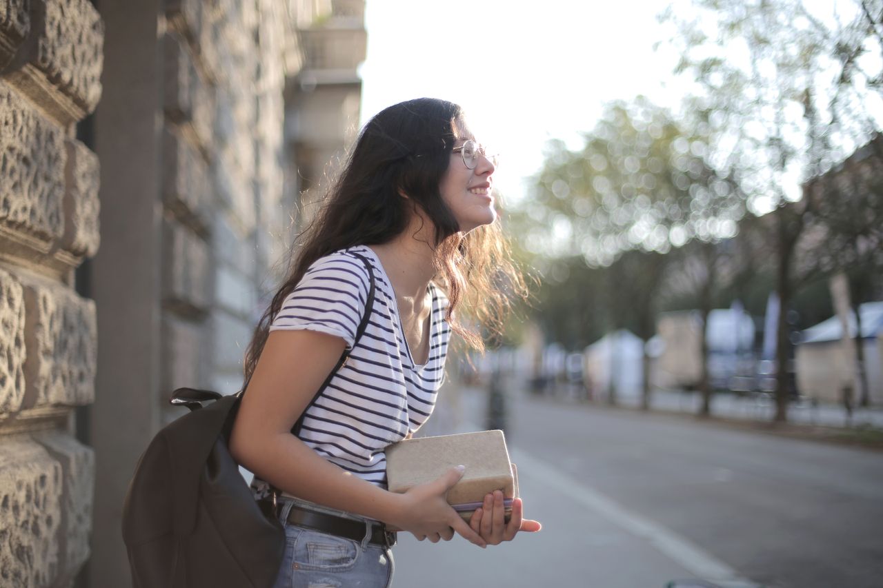 About Side view of smiling ethnic female in casual clothing standing with books and backpack on street near stone wall of building while chilling during weekend and looking away