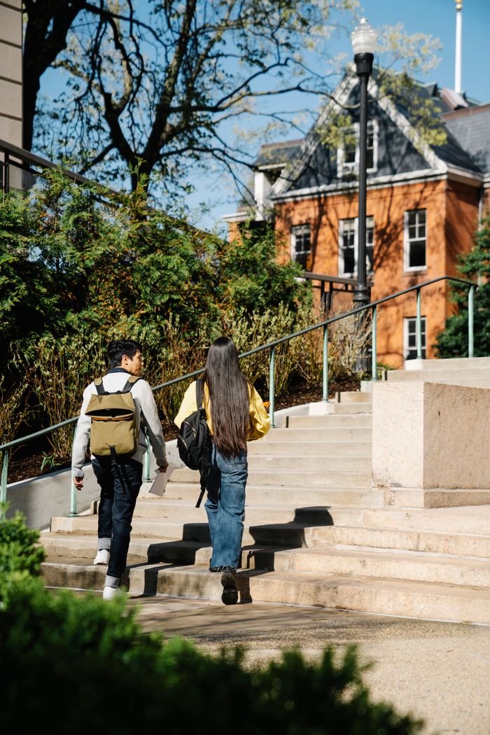 Home Two young adults walking up outdoor stairs surrounded by greenery and buildings.