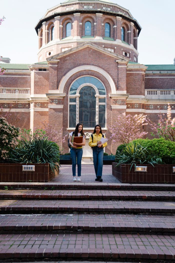 About Two young women walking past a historic university building on a sunny day.