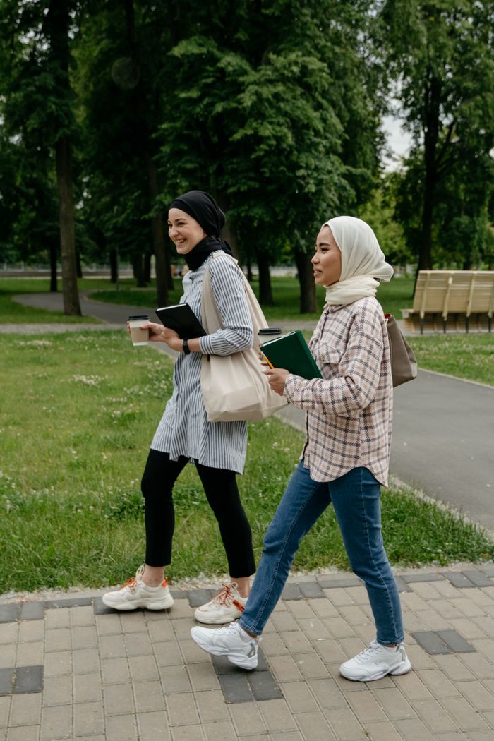 Two young Muslim women in hijabs walk through a green park, carrying books and coffee.